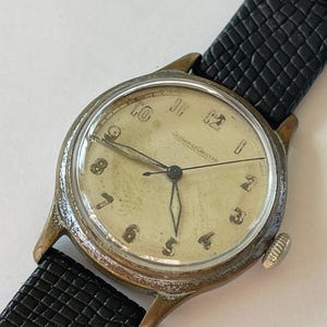 May include: Vintage Jaeger-LeCoultre wristwatch with a cream-colored face and black leather band. The watch has gold-toned hands and hour markers, with the brand name printed at the top. The black band is textured.