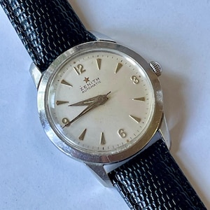May include: A silver Zenith Automatic wristwatch with a white face and black leather band. The watch face has black Roman numeral hour markers and a small star above the Zenith logo.
