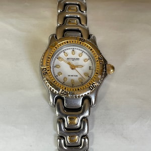 May include: A Wittnauer wristwatch with a silver and gold-tone metal band and bezel. The watch face is white with gold-tone hands and markers. The watch has a date display at the 3 o'clock position. The watch is approximately 1.25 inches in diameter.