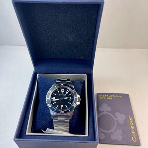 May include: A stainless steel Cortebert watch with a black dial and blue bezel, presented in a blue box. The watch features white hour markers and hands, with a date display. An instruction booklet is included.