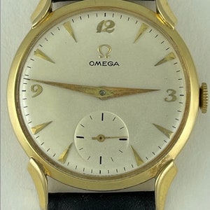 Pode incluir: Um relógio de pulso Omega dourado com um mostrador branco e uma pulseira de couro preta. O relógio tem marcadores de hora em algarismos romanos pretos e um pequeno mostrador de segundos.