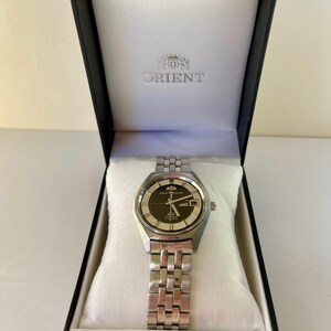 May include: A silver-toned Orient watch with a black dial and date display, presented in a black box with the Orient logo. The watch has a stainless steel band and is displayed on a white cushion.