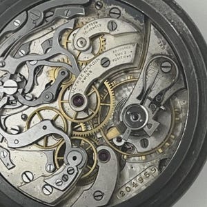 May include: The internal mechanical movement of a Ulysse Nardin pocket watch featuring gold-toned gears, silver-colored bridges, and levers. The metal plate is engraved with "Ulysse Nardin Swiss", "Fifteen 15 Jewels", "Adjusted Two 2 Positions", and "204959".