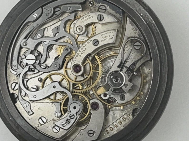 May include: The internal mechanical movement of a Ulysse Nardin pocket watch featuring gold-toned gears, silver-colored bridges, and levers. The metal plate is engraved with "Ulysse Nardin Swiss", "Fifteen 15 Jewels", "Adjusted Two 2 Positions", and "204959".