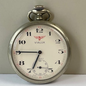 May include: Silver pocket watch with a white face, black hands, and black numerals. The watch face features the brand name "VIALUX" in red and a small seconds dial. The watch has a crown and a loop for a chain.