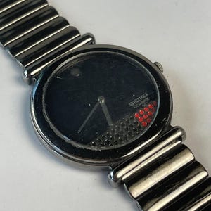 Puede incluir: Un reloj Seiko Quartz vintage con esfera negra y correa de metal plateado. La esfera presenta una pequeña sección de puntos rojos y el logotipo Seiko Quartz. La correa está hecha de eslabones metálicos acanalados y conectados.
