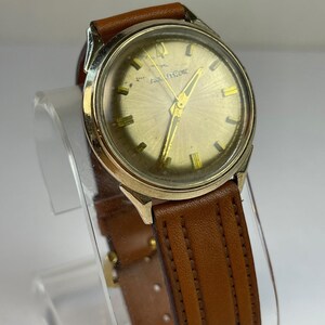 May include: A vintage wristwatch with a gold-toned face and hands, featuring the brand name "AQUATOR". The watch has a brown leather strap. The watch is displayed on a clear stand.