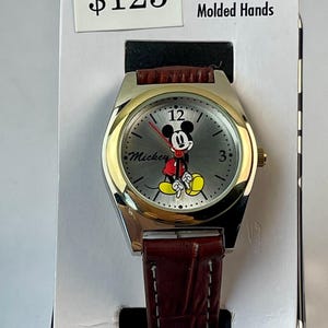 Pode incluir: Um relógio Mickey Mouse prateado e dourado com uma pulseira de couro castanho. O mostrador do relógio apresenta o Mickey Mouse com sua roupa clássica. O relógio está em sua embalagem original, que inclui o texto "Movable Molded Hands".