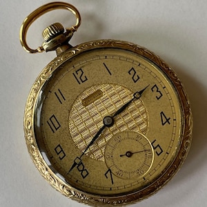 Puede incluir: Un reloj de bolsillo dorado con una esfera texturizada y números negros. El reloj presenta un pequeño dial de segundos y detalles ornamentados alrededor del bisel. Tiene un asa dorada y una corona texturizada.