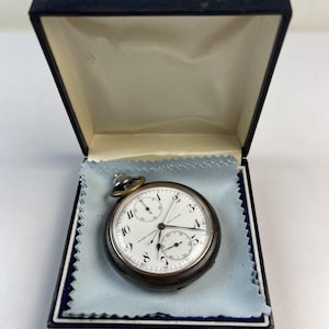 May include: A round Ulysse Nardin pocket watch with a dark metallic case and a white dial featuring black Arabic numerals, two sub-dials, and thin black hands. The watch rests on a light blue cloth inside a dark blue hinged presentation box.
