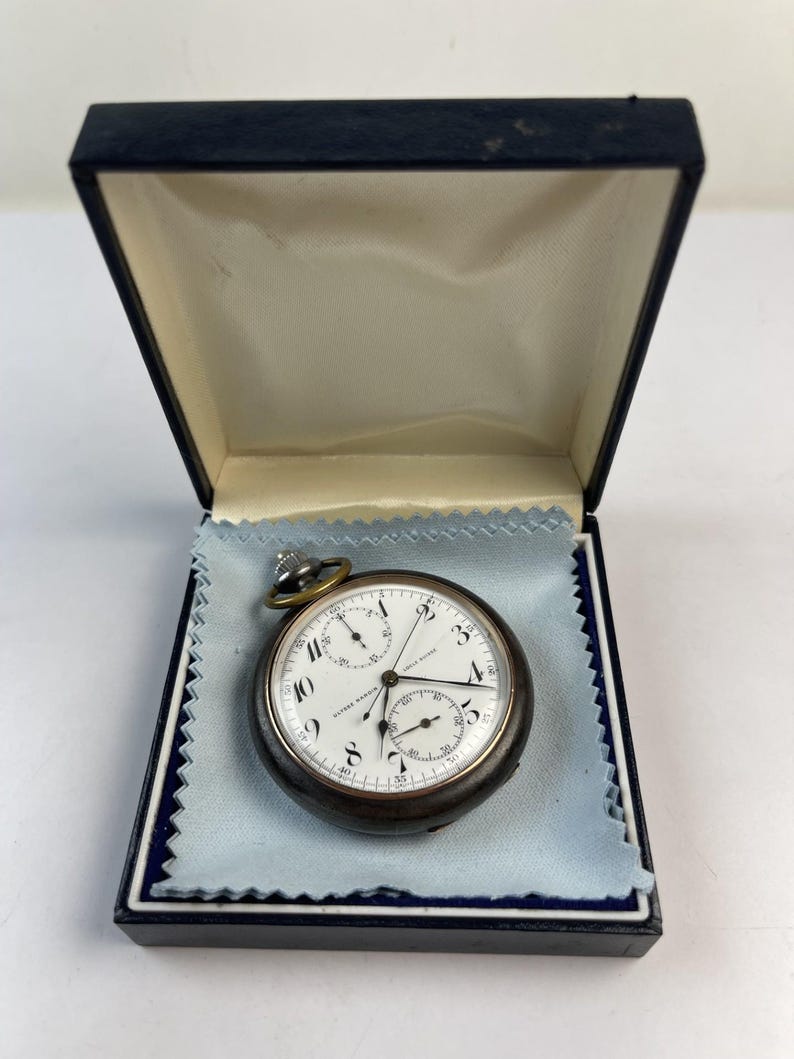 May include: A round Ulysse Nardin pocket watch with a dark metallic case and a white dial featuring black Arabic numerals, two sub-dials, and thin black hands. The watch rests on a light blue cloth inside a dark blue hinged presentation box.