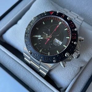 May include: A black and silver chronograph wristwatch with a red bezel and a black face. The watch has a date window and a day of the week window. The watch is in a black box.