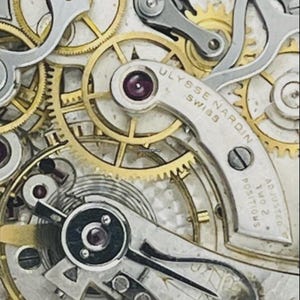 May include: A mechanical watch movement featuring gold-toned gears and silver-toned bridges. The bridge is engraved with ULYSSE NARDIN SWISS and ADJUSTED TWO 2 POSITIONS. The mechanism includes visible ruby jewels and a balance wheel assembly.