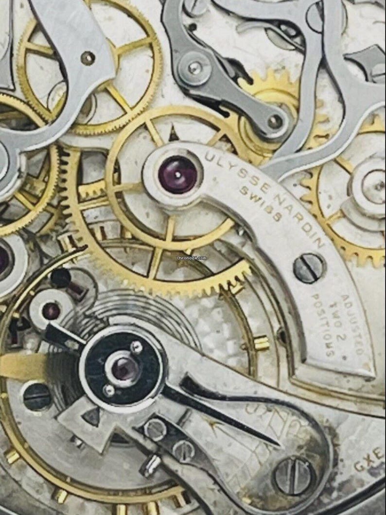 May include: A mechanical watch movement featuring gold-toned gears and silver-toned bridges. The bridge is engraved with ULYSSE NARDIN SWISS and ADJUSTED TWO 2 POSITIONS. The mechanism includes visible ruby jewels and a balance wheel assembly.