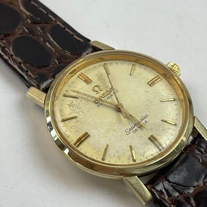 May include: A vintage gold-toned Omega Seamaster De Ville automatic wristwatch. The watch features a cream-colored face with gold hands and markers, and a brown leather band. The watch face displays the Omega logo and the words "Seamaster De Ville".