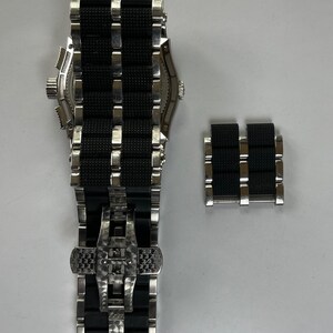 May include: A black and silver watch with a black rubber strap and a deployment clasp. The watch is shown with an extra strap link.