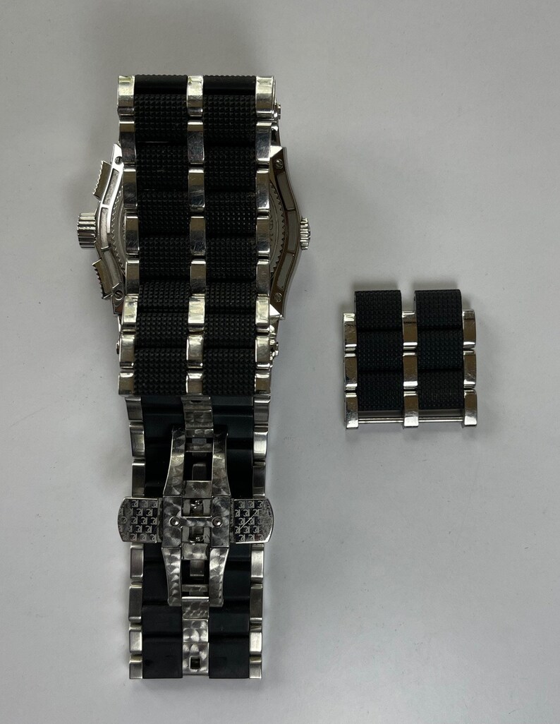 May include: A black and silver watch with a black rubber strap and a deployment clasp. The watch is shown with an extra strap link.