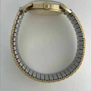 May include: A gold-tone wristwatch with a silver expandable band. The watch has a round face and a simple design.