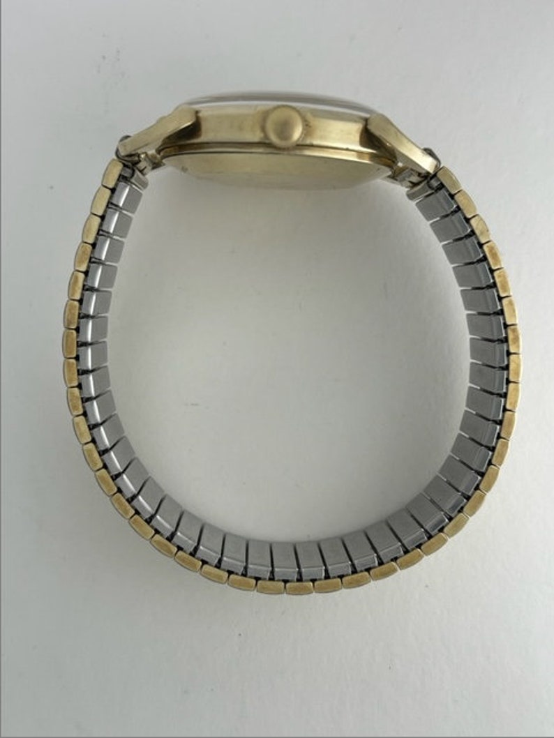 May include: A gold-tone wristwatch with a silver expandable band. The watch has a round face and a simple design.