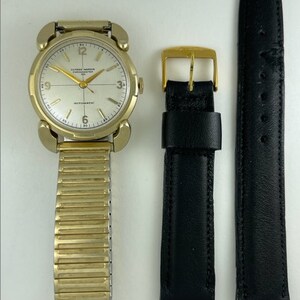 May include: A gold-toned wristwatch with a white face and black hands. The watch has a gold-toned metal band and a black leather strap. The watch face has the text "ULYSSE NARDIN CHRONOMETER" and "AUTOMATIC" printed on it.