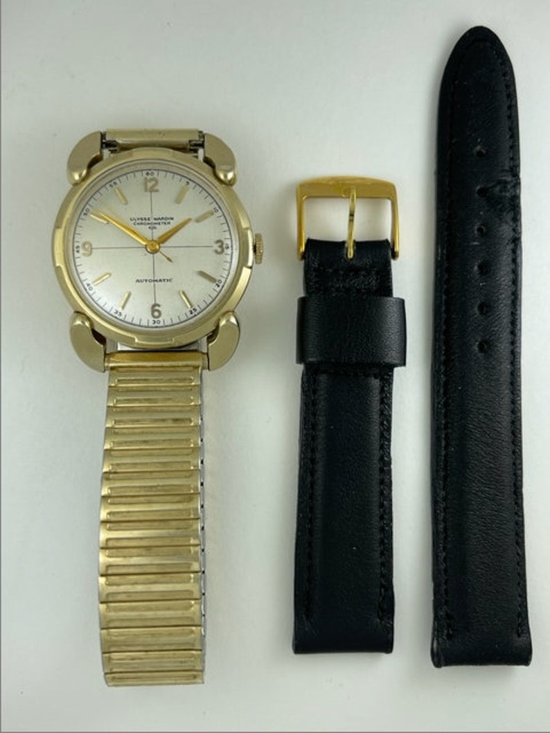 May include: A gold-toned wristwatch with a white face and black hands. The watch has a gold-toned metal band and a black leather strap. The watch face has the text "ULYSSE NARDIN CHRONOMETER" and "AUTOMATIC" printed on it.