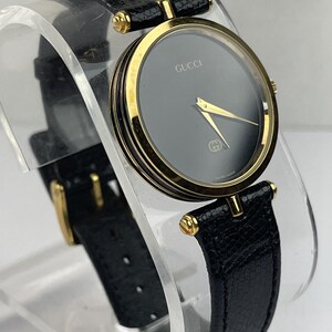 May include: A Gucci wristwatch featuring a round gold-tone case and a solid black dial with gold-tone hands. The watch includes a textured black leather strap with a gold-tone buckle. The brand name Gucci is printed in gold on the dial face.