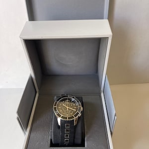 May include: A black and silver chronograph wristwatch with a black ceramic bezel and a black rubber strap with silver accents. The watch is in a white box with a gray interior.