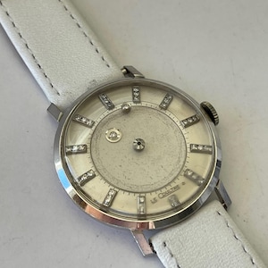 May include: A silver Le Coultre wristwatch with a white leather band. The watch face is surrounded by a row of diamonds.