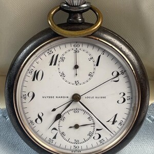 May include: An Ulysse Nardin Locle Suisse pocket watch with a dark metal case, a white dial featuring black Arabic numerals, two sub-dials, and a silver-toned crown with a brass-colored bow at the top. The watch face displays the brand name.