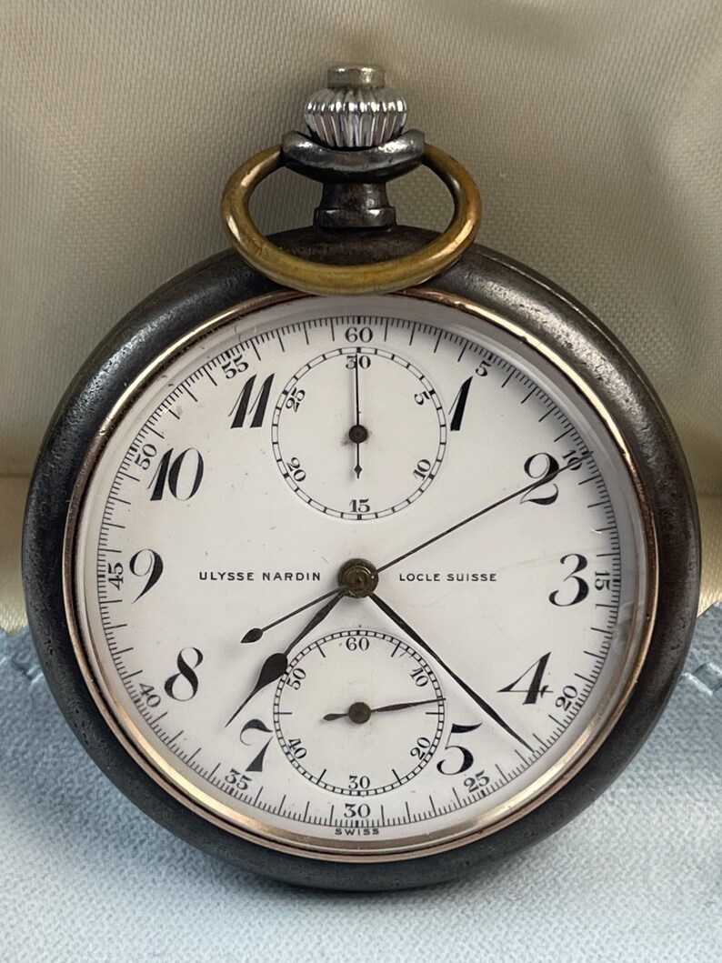 May include: An Ulysse Nardin Locle Suisse pocket watch with a dark metal case, a white dial featuring black Arabic numerals, two sub-dials, and a silver-toned crown with a brass-colored bow at the top. The watch face displays the brand name.