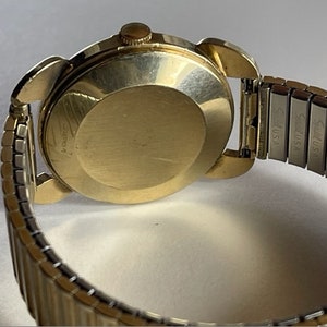 May include: A gold-tone wristwatch with a round face and a metal band. The watch has a stainless steel band with the text "Seiko USA" engraved on it.