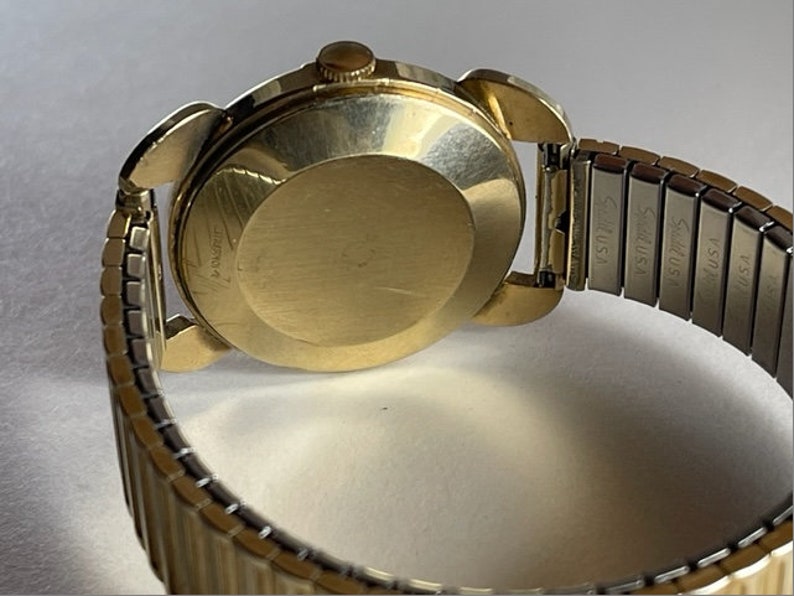 May include: A gold-tone wristwatch with a round face and a metal band. The watch has a stainless steel band with the text "Seiko USA" engraved on it.