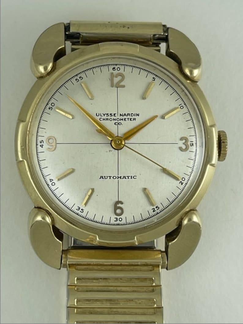May include: A gold-toned wristwatch with a white face and black hands. The watch face has the text "Ulysse Nardin Chronometer Co." and "Automatic".