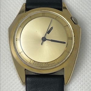 May include: A gold-toned Zodiac Olympos wristwatch with a black leather band. The watch face is gold with black hands and Roman numeral hour markers.