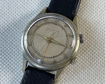 Puede incluir: Un reloj de pulsera plateado con una esfera blanca y una correa de cuero negra. La esfera del reloj tiene marcas negras y el texto "BENRUS WRISTALARM" en negro.