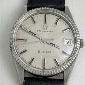 Puede incluir: Un reloj de pulsera Eterna-Matic 5 Star plateado con una correa de cuero negro. La esfera del reloj tiene marcadores de hora plateados y una ventana de fecha en la posición de las 3 en punto.