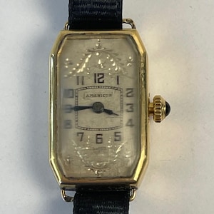 Op de afbeelding: Een vintage goudkleurig polshorloge met een rechthoekige wijzerplaat en een zwarte stoffen band. De wijzerplaat is wit met zwarte cijfers en wijzers, en het woord "AMERICUS" is zichtbaar. Het horloge heeft een kleine, zwarte, ronde kroon.