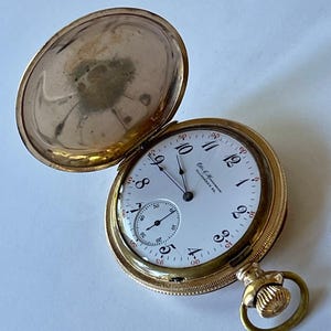 May include: A gold pocket watch with a white face and black hands. The watch has a small second hand dial at the 6 o'clock position. The watch face reads "C.H. Edmunds Allegheny, Pa."