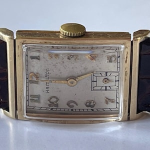 May include: A gold-toned wristwatch with a white face and black Roman numeral hour markers. The watch has a brown leather band and the word "HAMILTON" is printed on the face.