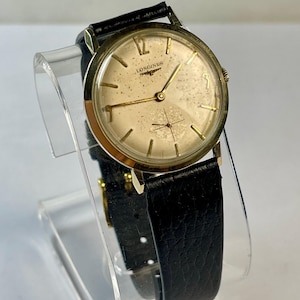 May include: A vintage Longines wristwatch with a gold-toned face and hands, featuring the brand name. The watch has a black leather band and a gold-colored case. The watch is displayed on a clear acrylic stand.