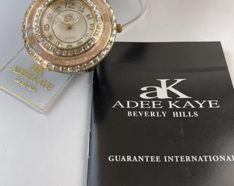 Könnte beinhalten: Eine runde Adee Kaye Beverly Hills Armbanduhr mit einer roségoldfarbenen, mit klaren Kristallen besetzten Lünette, einem weißen Zifferblatt mit AK-Logo und einem schwarzen, strukturierten Lederarmband. Inklusive passendem Etikett und Garantieheft.