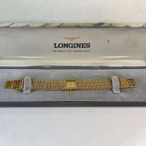 May include: A gold-toned Longines watch with a rectangular face and a matching gold band, presented in its original box. The box is gray with the Longines logo and text. The watch is displayed on a gray velvet-like surface.