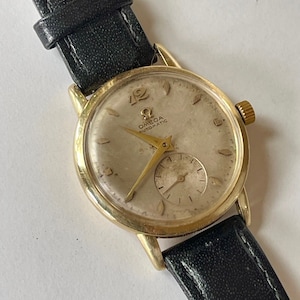 Könnte beinhalten: Eine goldfarbene Armbanduhr mit einem schwarzen Lederarmband. Das Zifferblatt der Uhr ist weiß mit schwarzen römischen Ziffern für die Stunden und einem kleinen Sekundenzeiger. Auf dem Zifferblatt der Uhr steht auch der Text "Omega Automatic".