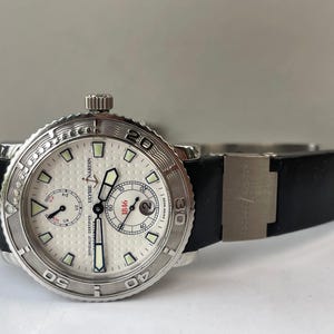May include: A silver-toned Ulysse Nardin watch with a white face and black band. The watch features a textured bezel, multiple sub-dials, and the text "OFFICIALLY CERTIFIED" and "1846". The band is black.