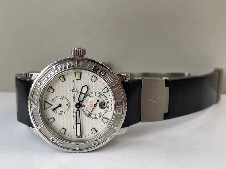 May include: A silver-toned Ulysse Nardin watch with a white face and black band. The watch features a textured bezel, multiple sub-dials, and the text "OFFICIALLY CERTIFIED" and "1846". The band is black.