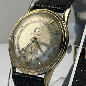 May include: A vintage Benrus wristwatch with a gold-tone round case and a textured off-white dial. The watch features gold-tone hour markers, a date indicator ring with numbers 1-31, a sub-dial, and a black textured leather strap with a buckle.