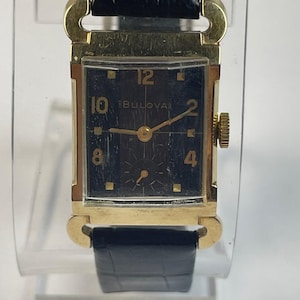 Puede incluir: Un reloj de pulsera Bulova vintage con una caja rectangular dorada y una esfera negra. El reloj presenta agujas e indicadores de hora dorados, con el logotipo de Bulova visible. El reloj tiene una correa de cuero negro.