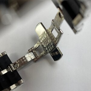 May include: Close-up of a silver watch band clasp with a black rubber strap. The clasp is engraved with "STAINLESS STEEL".