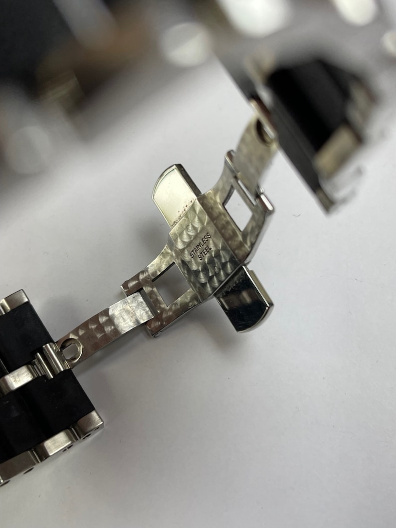 May include: Close-up of a silver watch band clasp with a black rubber strap. The clasp is engraved with "STAINLESS STEEL".