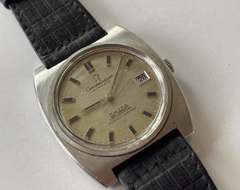 Puede incluir: Un reloj de pulsera Omega Constellation Automatic Chronometer Officially Certified de plata con una correa de cuero negro. La esfera del reloj tiene un fondo plateado con marcas negras y una ventana de fecha en la posición de las 3 en punto.